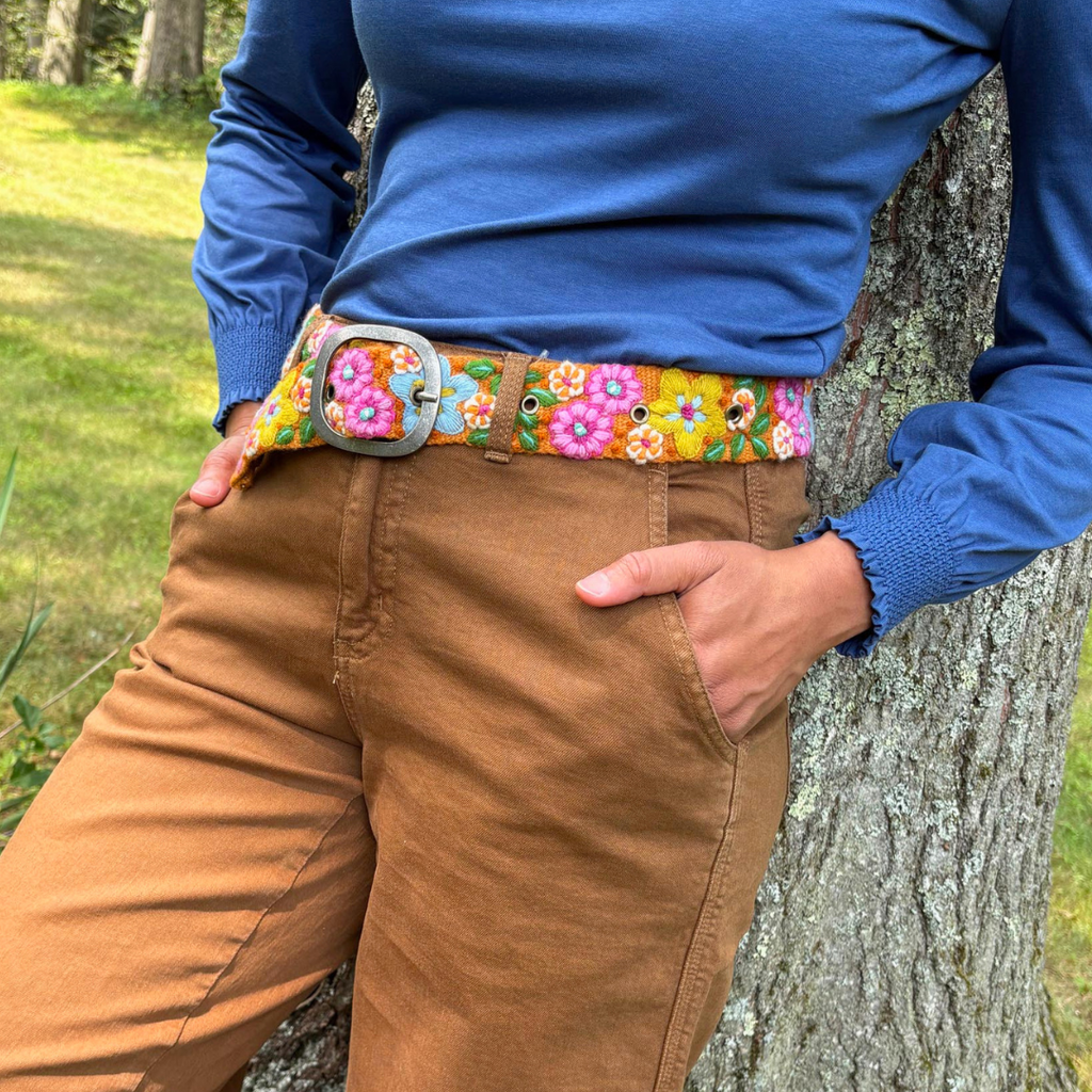 Person wearing a blue shirt and brown pants with a colorful belt, leaning against a tree.